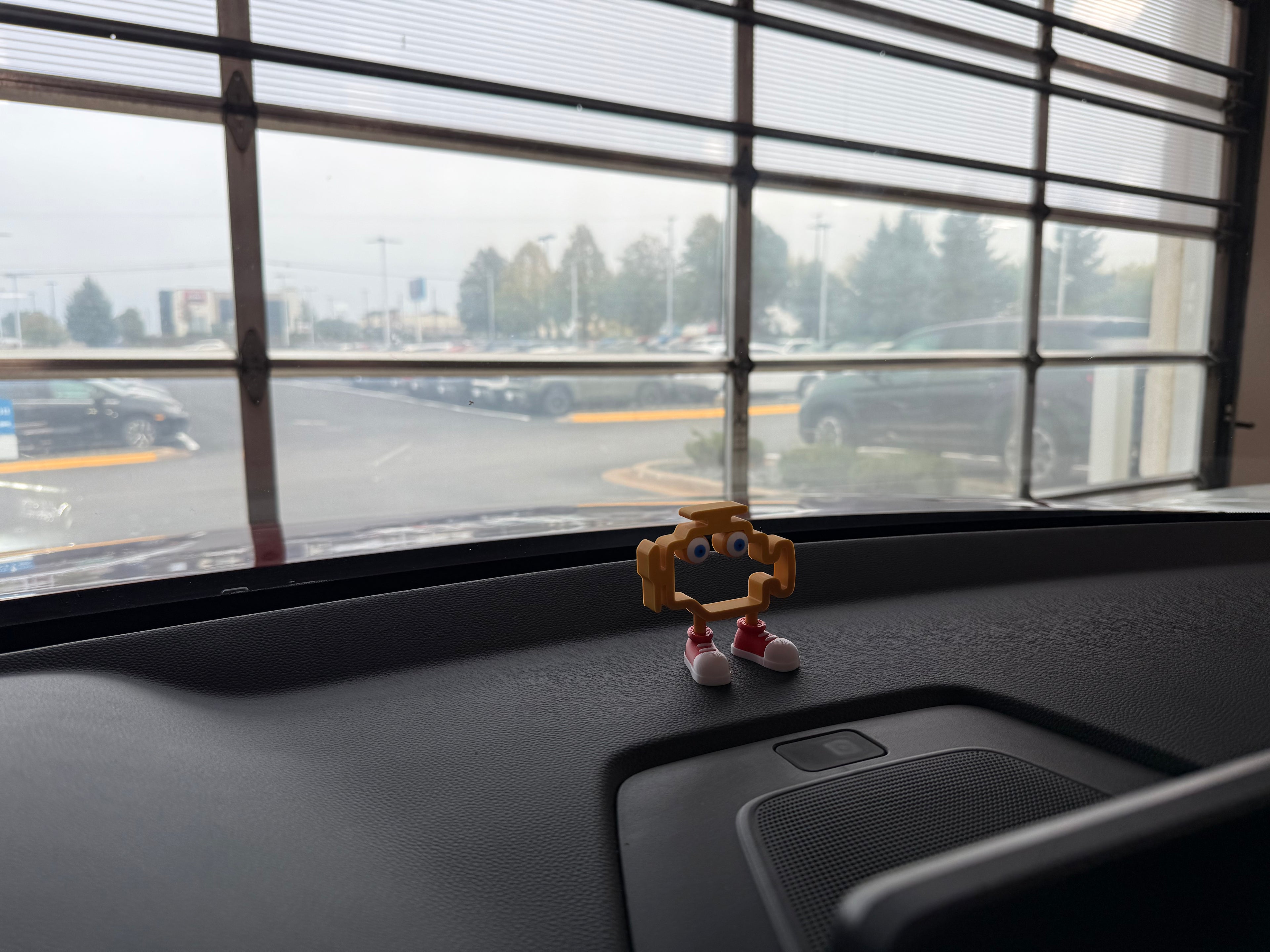 Small figurine on a car dashboard with a view of a parking lot through a window.