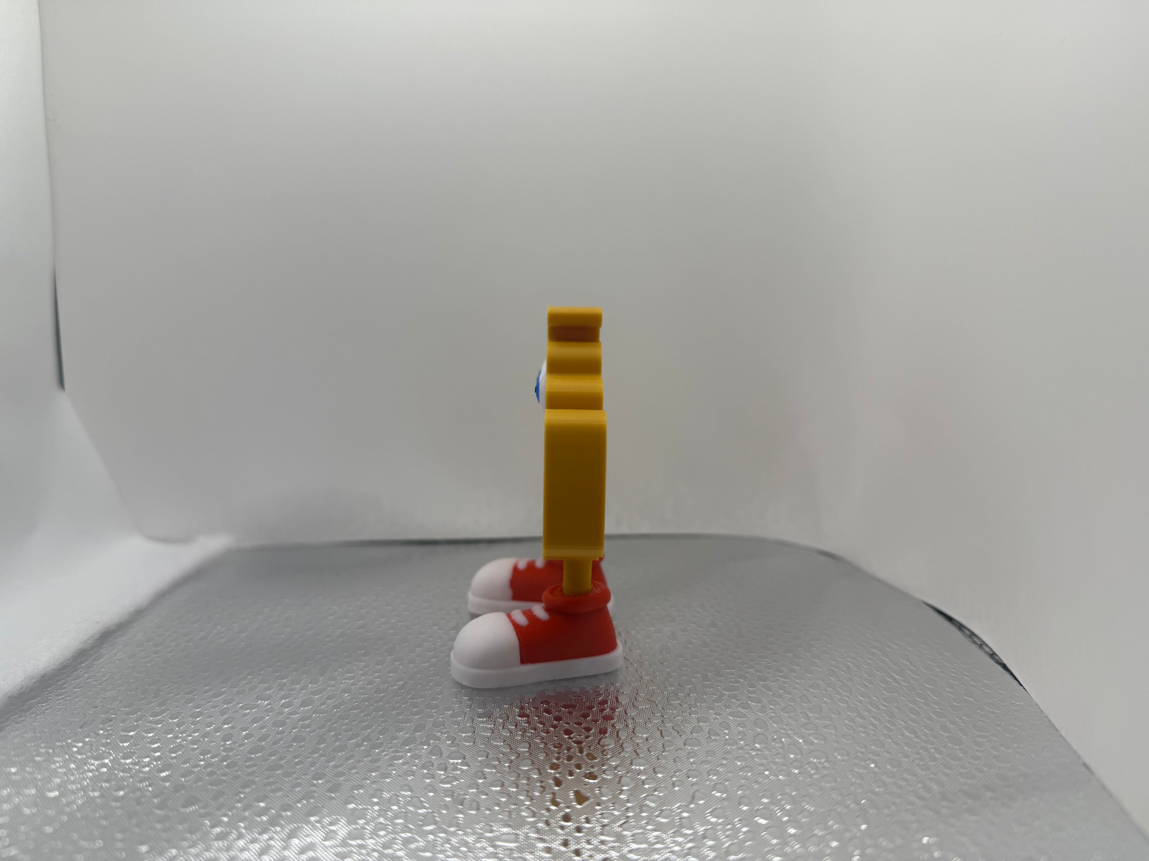 Small yellow toy figure with red shoes and a yellow block on a reflective surface.