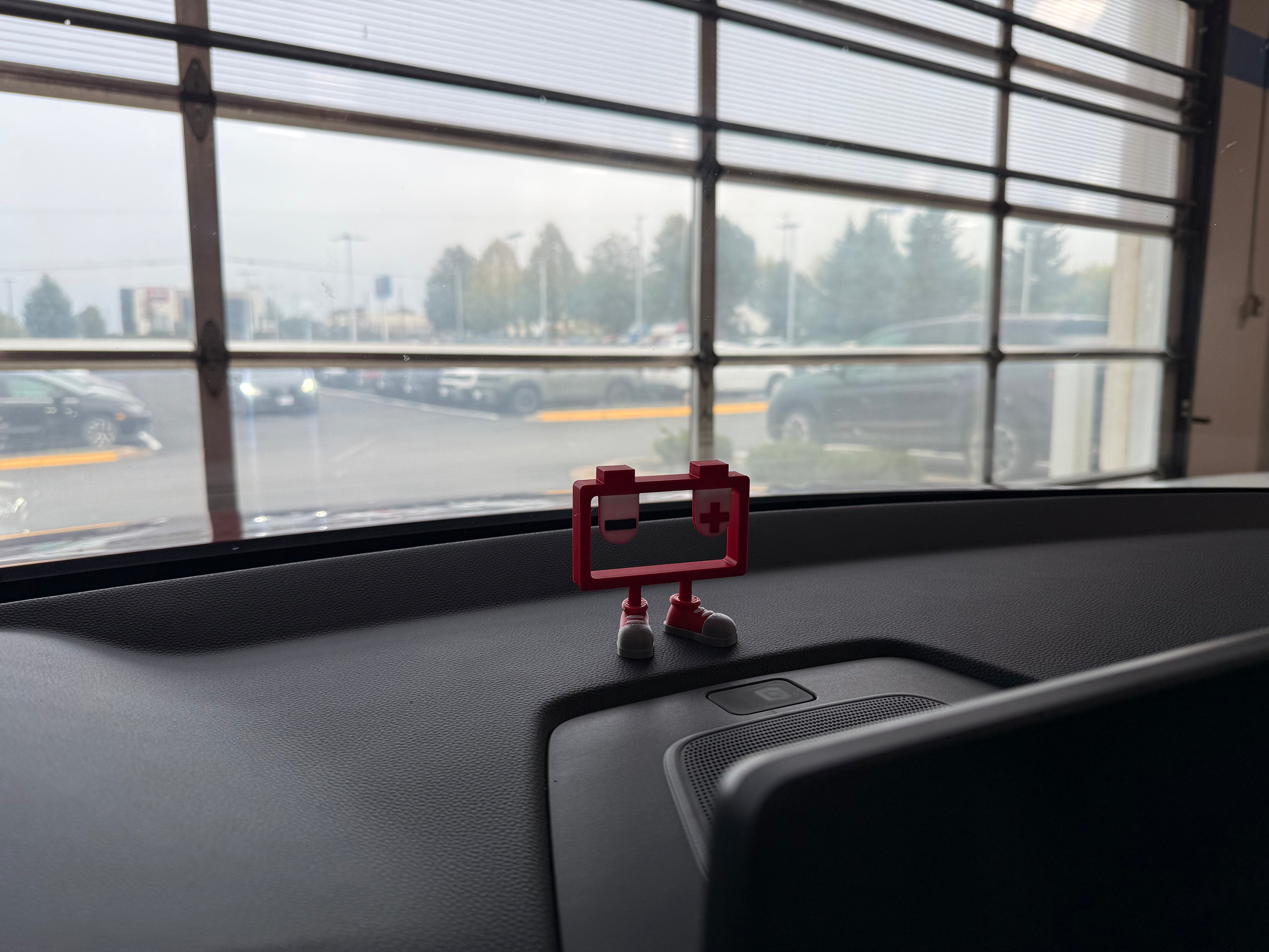 Small red robot figurine on a car dashboard with a view of a parking lot through the window.
