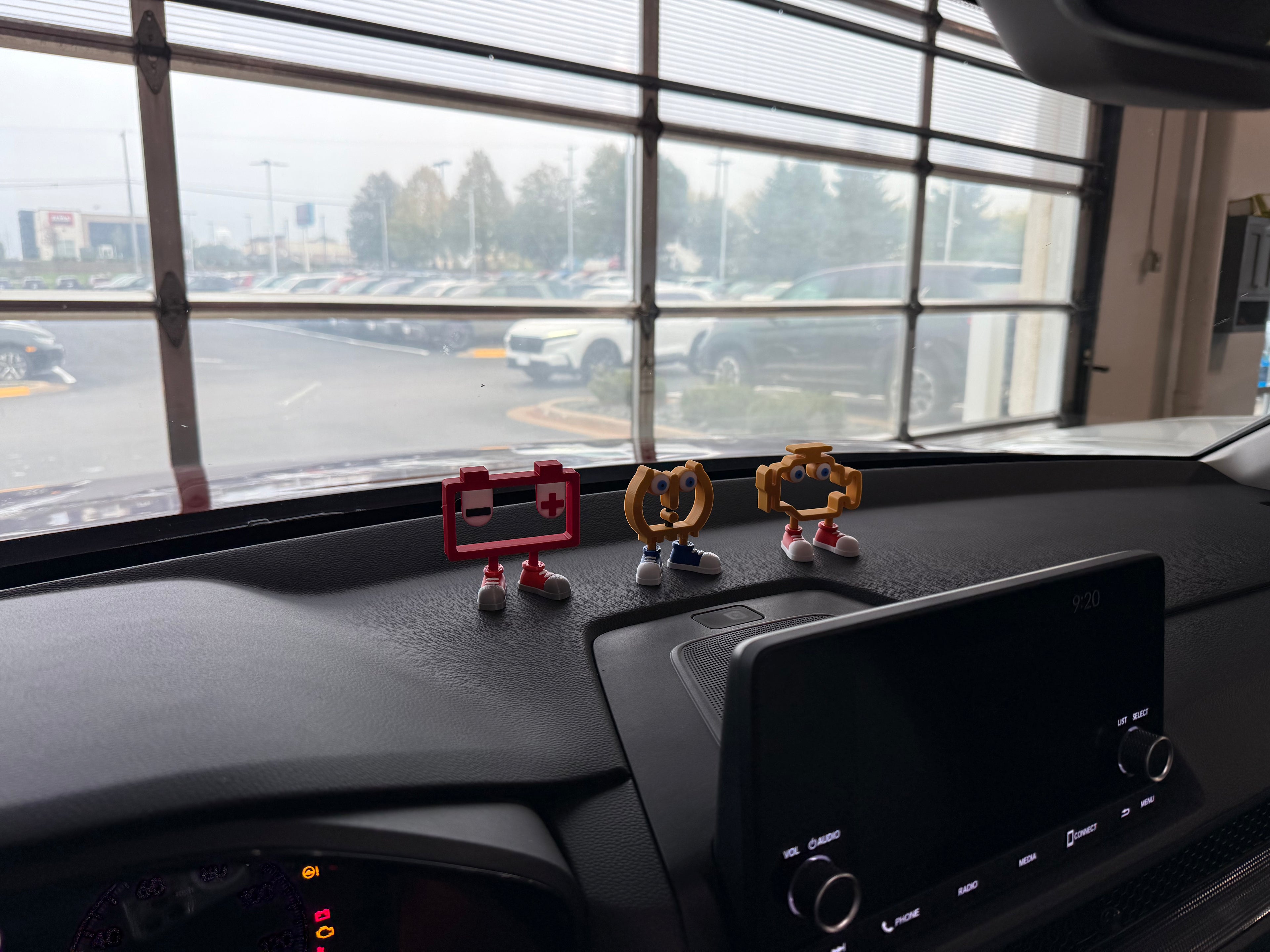 Car dashboard with figurines and a view of a parking lot through large windows.